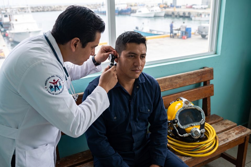 Médico MEDLAB realiza otoscopía a un buzo comercial junto a su casco de buceo de superficie